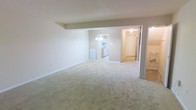 This two-floor residential space offers a practical and well-connected layout designed for comfortable everyday living. The main living area is generously sized, featuring wall-to-wall carpeting in a warm neutral tone that contributes to a clean, understated aesthetic throughout. Smooth white walls and standard ceilings provide a bright, airy feel, while recessed structural elements add subtle architectural definition to the open floor plan.

The living space flows naturally toward the kitchen, which is visible through an open passageway. The kitchen is equipped with white appliances, including a range and oven, and features wooden cabinetry along with a chandelier-style light fixture that adds a modest decorative touch. A wall-mounted heating unit is conveniently positioned in the main living area, ensuring year-round comfort.

Adjacent hallways lead to additional rooms across both floors, offering flexible use of space for sleeping, storage, or other needs. The overall color palette consists of soft beiges and whites, creating a cohesive and neutral environment that is easy to personalize. The layout is efficient and thoughtfully arranged, making good use of the available square footage across both levels. This space is well-suited for those seeking a clean, move-in-ready home with a straightforward and functional design.