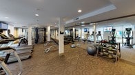 This fitness center offers a comprehensive workout environment designed to accommodate a wide range of exercise routines. The space is laid out across a single open floor, with patterned carpeting underfoot and a clean white ceiling fitted with recessed lighting that keeps the area well-illuminated throughout. Structural columns divide the floor plan into functional zones while full-length mirrors along the walls visually expand the space and allow users to monitor their form during exercise. Large windows along one side of the room invite natural light and provide views of the surrounding greenery, creating an energizing atmosphere. The cardio zone is well-stocked with Matrix-brand treadmills, elliptical trainers, and stationary bikes, each equipped with individual display screens. Wall-mounted flat-screen televisions are positioned for easy viewing from multiple areas of the room. A multi-functional cable machine anchors the strength training section, complemented by a dedicated free weights area featuring a full rack of dumbbells in graduated sizes. Additional training accessories include stability balls, a BOSU balance trainer, kettlebells, and a flat weight bench. A mirrored wall along the free weights section further enhances the sense of openness. Access to the fitness center is provided through glass-paneled doors, and a small storage rack near the entrance holds additional equipment and supplies.