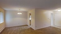This single-floor residence offers a clean, functional layout with a warm and neutral aesthetic throughout. The main living and dining area is generously sized, featuring soft beige carpet that adds a sense of warmth underfoot. A classic chandelier hangs overhead, providing focused ambient lighting and serving as a subtle decorative focal point in the dining zone. The walls are finished in a light, neutral tone that keeps the space feeling bright and open. A partial wall division separates the main living area from the kitchen, which is accessible through a doorway. The kitchen features warm wood cabinetry, white appliances, and a compact, efficient layout. The cabinetry extends to the upper portion of the walls, maximizing storage capacity. White baseboards throughout provide a clean contrast to the carpeted floors, contributing to the overall tidy and well-maintained appearance of the space. The single-floor design ensures easy accessibility and a cohesive flow between areas. Overall, the space presents a practical and comfortable living environment with a straightforward, classic interior style suited for everyday living.