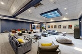 Located at 99 Gold Star Hwy in Groton, Connecticut, this single-floor facility offers a generously sized, multipurpose recreational environment designed for both leisure and social engagement. The interior is unified by a consistent design palette of navy blue accents, neutral carpeting, and drop-tile ceilings with recessed lighting throughout.

The main gathering area features high-top bar tables and stools, motivational artwork, and an American flag display, creating a welcoming communal atmosphere. Adjacent to this is a standout recreational zone housing an indoor putting green with multiple flags and a cornhole board, surrounded by large windows that flood the space with natural light and offer views of the surrounding greenery. A distinctive arched decorative window adds architectural character to this room.

A dedicated media lounge area showcases plush sofas, ottomans, a wall-mounted television, and a striking recessed skylight feature in the ceiling, lending the space an upscale, comfortable ambiance. Movie posters line the walls, enhancing the entertainment-focused character of the room.

The entryway is particularly dramatic, featuring exposed brick walls, navy-painted columns, and a grand crystal chandelier that sets an elevated tone upon arrival. A fitness area and additional seating nooks with open shelving complete the layout, making this a well-rounded, activity-rich facility.