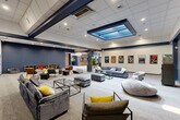 Located at 99 Gold Star Hwy in Groton, Connecticut, this single-floor facility offers a generously sized, multipurpose recreational environment designed for both leisure and social engagement. The interior is unified by a consistent design palette of navy blue accents, neutral carpeting, and drop-tile ceilings with recessed lighting throughout.

The main gathering area features high-top bar tables and stools, motivational artwork, and an American flag display, creating a welcoming communal atmosphere. Adjacent to this is a standout recreational zone housing an indoor putting green with multiple flags and a cornhole board, surrounded by large windows that flood the space with natural light and offer views of the surrounding greenery. A distinctive arched decorative window adds architectural character to this room.

A dedicated media lounge area showcases plush sofas, ottomans, a wall-mounted television, and a striking recessed skylight feature in the ceiling, lending the space an upscale, comfortable ambiance. Movie posters line the walls, enhancing the entertainment-focused character of the room.

The entryway is particularly dramatic, featuring exposed brick walls, navy-painted columns, and a grand crystal chandelier that sets an elevated tone upon arrival. A fitness area and additional seating nooks with open shelving complete the layout, making this a well-rounded, activity-rich facility.