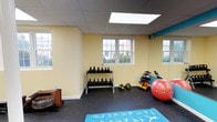 This yoga and fitness room is a thoughtfully designed multipurpose exercise space finished with durable rubber flooring and soft yellow walls that create an energizing yet welcoming atmosphere. A dropped tile ceiling features multiple recessed skylights that flood the room with bright, even illumination, complementing the natural light that streams in through several double-hung windows offering views of the surrounding exterior landscape.

A full-length wall mirror runs along one side of the room, visually expanding the space and supporting proper form during workouts. Vibrant teal accent trim adds a bold splash of color against the neutral walls, reinforcing the room's active character. The open floor plan accommodates yoga mats laid out for stretching and mat-based exercises, while a dedicated equipment zone houses a tiered kettlebell rack, a colorful dumbbell rack, medicine balls, and a stability ball.

A WaterRower Club rowing machine occupies a quieter corner of the room, ideal for low-impact cardio training. Additional amenities include an adjustable weight bench and a structural column integrated into the layout. Two interior doors provide access to adjoining areas, ensuring smooth circulation throughout the space.