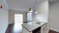 Unit 410 at 333 Central Avenue is a single-floor residence designed with an open-concept layout that seamlessly connects the kitchen and living areas. The kitchen serves as a central hub, anchored by a peninsula island topped with light granite-style countertops and an undermount stainless steel sink. Two elegant pendant lights with gold-toned shades hang above the island, providing both functional task lighting and a refined aesthetic accent. White cabinetry and a built-in dishwasher complete the kitchen's clean, contemporary look. The living area flows naturally from the kitchen and is filled with natural light, courtesy of large sliding glass doors and an adjacent window fitted with horizontal blinds. The sliding doors open to an outdoor balcony, offering views of the surrounding landscape. Rich hardwood flooring runs throughout the main living space, adding warmth and texture to the neutral color palette of soft gray and white walls. Crown molding along the ceiling provides a polished architectural detail that elevates the overall finish of the unit. The result is a bright, functional, and well-appointed space ideal for both everyday living and entertaining.