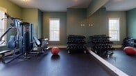 This fitness center is a comprehensively outfitted exercise space designed to support a full range of workout routines. The room is finished with distinctive sage green walls and durable dark rubber flooring, which provides both traction and impact absorption during exercise. Natural light filters in through multiple windows fitted with blinds, complementing the flush-mounted ceiling fixtures that ensure consistent illumination throughout.

The space is organized into functional zones. A dedicated cardio area houses two treadmills and an elliptical trainer, accompanied by a wall-mounted flat-screen television for entertainment during workouts. The strength training section features a multi-function cable and weight machine along with an adjustable bench, catering to a variety of resistance exercises. A well-stocked free weight area includes a tiered dumbbell rack holding a broad range of weights, alongside a medicine ball for functional training.

Full-length mirrors line one wall, visually expanding the space while allowing users to monitor their form. Entry is through a glass-paneled door clearly marked as the Fitness Center, connecting to an adjacent common area. The overall layout is practical and well-organized, offering an accessible, all-in-one exercise environment.