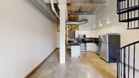 Located at 1900 E Superior Ave in Cleveland's vibrant Superior district, this two-story loft unit offers a compelling blend of historic industrial character and contemporary upgrades. The main level features polished concrete tile flooring, soaring ceilings with exposed wooden beams, visible ductwork, and a striking black iron spiral staircase that serves as a focal point of the open living area. Large grid-pane windows allow natural light to fill the space. The well-appointed kitchen showcases white cabinetry, dark granite countertops, a mosaic tile backsplash, stainless steel appliances including a range, microwave, and refrigerator, and track lighting overhead. The bathroom is finished with mosaic tile accents, a tub-shower combo, a white vanity with granite countertop, and industrial-style pendant lighting. The upper level features carpeted open rooms beneath a dramatic angled ceiling of exposed timber beams and original wood planking, lending a raw, loft-like atmosphere. Additional rooms on both levels offer warm-toned hardwood flooring, pendant lighting, and ample closet storage. The overall ambiance is one of urban industrial chic, combining the building's original architectural bones with thoughtful modern renovations.