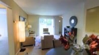 This single-floor space offers a welcoming and functional layout designed for comfortable everyday living. The living room serves as the heart of the home, featuring a generous seating arrangement with plush sofas and accent chairs arranged around a patterned area rug that anchors the space. Warm, neutral tones on the walls complement the light-colored flooring, creating a cohesive and inviting ambiance throughout.

A standout feature of the living area is the large sliding glass door that spans a significant portion of the exterior wall, allowing an abundance of natural light to flood the interior and offering a pleasant view of the lush greenery outside. Sheer curtains frame the door, softening the light while maintaining an airy, open feel.

The room is thoughtfully accessorized with decorative elements, including a distinctive round wall mirror, an open media console with styled décor, and coordinated accent lighting provided by a classic floor lamp. A ceiling fan is also present, adding both functionality and a casual, relaxed character to the space.

Overall, the space strikes a balance between practicality and style, with a bright, neutral palette and well-chosen furnishings that create a comfortable and visually cohesive environment suited for both relaxing and entertaining.