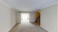 This two-story residence offers a well-organized layout spread across two floors, providing ample space for comfortable everyday living. The main level features a generously sized living area finished with plush wall-to-wall carpeting in a warm neutral tone, creating an inviting and cozy atmosphere throughout. Crown molding accents the ceilings, adding a refined architectural detail that complements the clean, neutral color palette of the walls and trim.

An open passage connects the main living area to an adjoining room, which benefits from abundant natural light streaming through a set of French doors — lending brightness and an airy quality to the space. The layout encourages an easy flow between rooms, making the interior feel open and connected.

A classic staircase with a warm-toned wood banister and turned spindles anchors one side of the main floor, providing an elegant transition to the upper level. The staircase serves as a subtle focal point, contributing character and traditional charm to the interior.

Throughout the space, the consistent use of neutral tones, soft carpeting, and classic trim work creates a cohesive and understated aesthetic that is both functional and welcoming — well-suited to a variety of furnishing styles and personal tastes.