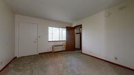 This single-floor unit offers a practical and straightforward layout with a warm, classic character defined by its distinctive wood-paneled accent walls. The kitchen is efficiently designed as a galley-style space, featuring warm wood cabinetry, light stone-finish countertops with a stainless sink, and white appliances including a range and refrigerator, all set on easy-care laminate hardwood flooring. A striking floor-to-ceiling wood-paneled partition wall separates the kitchen area from the main living space, adding a natural, rustic element to the interior. The living area is open and generously proportioned, with carpeted flooring, neutral-toned walls, and a window that brings in natural light and views of the surrounding greenery. A wall-mounted HVAC unit provides climate control. The bedroom is a calm, carpeted retreat featuring a large sliding window that frames outdoor views and fills the room with ample natural light. The bathroom is clean and functional, equipped with a tub and shower combination, a white vanity with storage, and a mirrored medicine cabinet above, all finished with warm laminate wood flooring. Wood-tone doors and trim details carry a consistent, cohesive aesthetic throughout the entire space.