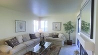 This single-floor residence offers a comfortable and well-designed living environment with a calm, neutral aesthetic that is both inviting and functional. The living room serves as the heart of the space, featuring plush upholstered sofas arranged around a glass-top coffee table that rests on a classic area rug. Soft, light-toned walls create a serene backdrop, complemented by framed botanical artwork that adds a refined decorative touch. Corner windows allow natural light to fill the room, enhancing its open and airy atmosphere. The high, smooth ceiling amplifies the sense of spaciousness, giving the room an elevated feel. A large indoor tree adds a vibrant natural element, introducing warmth and life to the corner of the room. The carpeted flooring contributes to the overall softness and comfort of the space. Subtle details throughout — including decorative pillows, a table lamp, and carefully placed accent pieces — reflect a thoughtful approach to interior styling. The overall ambiance is tranquil and well-maintained, making this a welcoming space suited for both relaxation and everyday living.