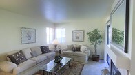 This single-floor residence offers a comfortable and well-designed living environment with a calm, neutral aesthetic that is both inviting and functional. The living room serves as the heart of the space, featuring plush upholstered sofas arranged around a glass-top coffee table that rests on a classic area rug. Soft, light-toned walls create a serene backdrop, complemented by framed botanical artwork that adds a refined decorative touch. Corner windows allow natural light to fill the room, enhancing its open and airy atmosphere. The high, smooth ceiling amplifies the sense of spaciousness, giving the room an elevated feel. A large indoor tree adds a vibrant natural element, introducing warmth and life to the corner of the room. The carpeted flooring contributes to the overall softness and comfort of the space. Subtle details throughout — including decorative pillows, a table lamp, and carefully placed accent pieces — reflect a thoughtful approach to interior styling. The overall ambiance is tranquil and well-maintained, making this a welcoming space suited for both relaxation and everyday living.