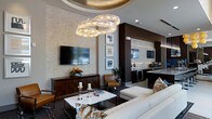 This elegantly designed single-floor space exudes a contemporary luxury aesthetic through a carefully curated combination of materials, lighting, and furnishings. The open-concept layout connects a generously sized lounge area to a fully equipped wet bar, creating a seamless flow ideal for entertaining.

The lounge area is anchored by a pair of striking ring-shaped crystal chandeliers suspended from a coffered ceiling, casting warm, ambient light across the space. A textured stone accent wall adds depth and visual interest, serving as a backdrop for a wall-mounted flat-screen television above a dark wood credenza. Seating arrangements include a plush white sectional sofa, patterned accent chairs, and a marble-topped coffee table, all positioned atop a bold geometric area rug.

The wet bar area features dark cabinetry with under-counter beverage refrigerators, a marble countertop, and a second wall-mounted television, making it a functional and stylish hub. Dark structural columns contrast with the lighter stone walls throughout, reinforcing the space's upscale, modern character. The overall ambiance is polished and inviting, with rich material contrasts contributing to a refined yet comfortable atmosphere.