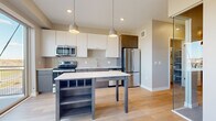 This single-floor residence showcases a clean, contemporary design with light hardwood flooring flowing throughout. The open-concept kitchen is a standout feature, equipped with stainless steel appliances including a refrigerator, range, microwave, and dishwasher. Two-tone cabinetry pairs white upper cabinets with warm wood-grain lower cabinets, complemented by a glossy subway tile backsplash and crisp white countertops. Pendant lighting hangs above a freestanding island with open shelving beneath, offering additional storage and prep space. Natural light pours in through large sliding glass doors that provide access to a balcony with exterior views.

A hallway connects the living areas to the private spaces, where a well-sized bedroom benefits from a large window framing outdoor views and a dedicated walk-in closet with built-in shelving. A glass partition adds a modern architectural touch near the bedroom entry. The bathroom features a soaking tub with tiled surround, a sleek vanity with wood-toned cabinetry, white countertop, and light hardwood-style flooring. The overall palette of warm neutrals and clean finishes creates a cohesive, understated aesthetic throughout the residence.