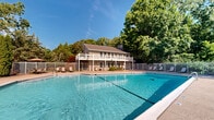 This outdoor aquatic facility offers a welcoming and well-maintained environment centered around a generously sized swimming pool with crystal-clear blue water. The pool is equipped with stainless steel entry ladders and features clearly marked depth indicators, including a designated no-diving zone, ensuring both safety and functionality for swimmers of all levels. The surrounding pool deck is finished in a textured, slip-resistant surface and is furnished with an array of lounge chairs and umbrellas, providing ample space for relaxation and sunbathing. The entire pool area is enclosed by a white metal fence, offering both security and a clean, classic aesthetic. Mature trees and lush greenery frame the perimeter, creating a sense of seclusion and natural tranquility while filtering natural light throughout the day. Anchoring the space is a two-story clubhouse building clad in neutral siding and crowned with a pitched roof. The structure features a covered upper-level balcony with white railings, offering an elevated vantage point over the pool area. Ground-level access doors open directly onto the pool deck, facilitating easy indoor-outdoor flow. Additional seating areas with tables and umbrellas are positioned along the deck, making this facility well-suited for both leisure and social gatherings.