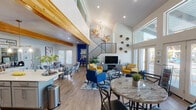 This vibrant two-story space is thoughtfully designed to accommodate a wide variety of activities and functions. The main level opens into a dramatic, soaring great room with vaulted ceilings, exposed natural wood beams, and light wood flooring throughout. A striking open staircase with metal railings connects the two floors, while large glass doors and expansive windows flood the space with natural light and provide direct views of an outdoor pool area.

The fully equipped kitchen is a standout feature, boasting grey shaker-style cabinetry, stainless steel appliances, a mosaic tile backsplash, a large central island with bar seating, and bold pendant lighting beneath the wood beam ceiling. Adjacent to the kitchen, a dining and lounge area offers a marble-top table and a comfortable seating arrangement with colorful accent chairs.

A dedicated yoga and fitness studio offers an open, serene environment complete with exercise balls, yoga mats, wall mirrors, and motivational artwork. The children's activity rooms are bright and imaginative, featuring small-scale tables and chairs, a large built-in bookshelf with colorful cubbies, whiteboards, and ample storage. A quiet workspace nook with pool-facing French doors rounds out this well-appointed, multi-functional space.