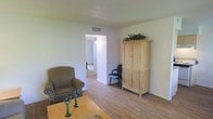 This single-floor space features a functional and straightforward layout that emphasizes practicality and comfort. The living area is the focal point of the space, showcasing light-toned hardwood-style flooring that runs throughout, lending warmth and continuity to the interior. The walls are finished in a neutral off-white palette, complemented by a textured ceiling that adds subtle visual interest. Natural and ambient light keeps the space feeling bright and airy.

A standout feature of the living area is a tall, light-wood armoire with cabinet doors, topped with decorative greenery that adds a natural accent. Seating is provided by a classic houndstooth-patterned armchair, contributing a touch of traditional character. A wooden coffee table anchors the seating arrangement, while a simple office-style chair occupies a corner near the armoire.

A central hallway or doorway connects the living area to an adjoining room, suggesting a multi-room floor plan with a logical and easy-to-navigate flow. The overall ambiance is clean, modest, and livable, making it a comfortable and unpretentious space suited for everyday use.