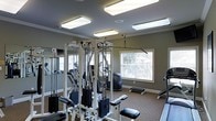 This single-floor fitness center is thoughtfully designed to provide a comprehensive exercise environment. The space is anchored by a multi-station cable and weight machine system, offering a wide range of strength training options suitable for users of varying fitness levels. Complementing the strength equipment, the room also houses cardio machines, including what appears to be a treadmill and an elliptical trainer, ensuring a well-rounded workout experience.

The interior is finished with neutral gray walls and light-toned carpeted flooring, creating a clean and focused atmosphere. Large windows along one wall allow natural light to filter into the space, while multiple flush-mounted rectangular overhead light fixtures ensure consistent, bright illumination throughout the room. Crown molding along the ceiling adds a subtle architectural touch that elevates the overall finish of the space.

Full-length mirrors are positioned along one wall, a practical feature that allows users to monitor their form during exercise while also enhancing the sense of openness. The layout is organized to allow comfortable movement between equipment, making the space both functional and accessible. Overall, the fitness center strikes a balance between utility and comfort, offering a well-maintained environment for daily physical activity.