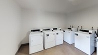 This shared laundry room is a practical, well-organized facility designed to serve multiple residents efficiently. The space is accessed through a glass-paneled door that allows natural light to filter into the entry area, which features neutral-toned walls, light flooring, a stainless steel waste bin, and a posted laundry room guidelines sign for user reference. A wall-mounted utility box is also present near the entrance.

The main laundry area is clean and straightforward in design, with bright white walls and smooth light-toned flooring throughout. The room accommodates multiple commercial-grade top-loading washing machines and front-loading dryers, all in white, arranged in an orderly configuration along the walls. The machines are equipped with digital displays and coin-operated controls, indicating shared-use functionality. Overhead lighting keeps the space well-lit, contributing to a clean and functional atmosphere.

The overall ambiance is utilitarian and well-maintained, prioritizing ease of use and efficiency. The clear layout allows users to move comfortably between machines, making laundry tasks straightforward and convenient.