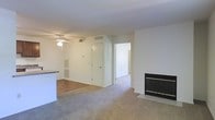 This single-floor space offers an open, functional layout designed for comfortable everyday living. The main living area is finished with plush carpeting and anchored by a sleek, black-framed fireplace set into a clean white wall, serving as a natural focal point. A ceiling fan overhead provides both light and air circulation throughout the space.

The layout flows seamlessly from the living area toward additional rooms, with a hallway visible in the background leading to what appears to be a bedroom or secondary space. A doorway nearby likely accesses a closet or utility area, contributing to practical storage options within the unit.

To one side, the living space transitions into a kitchen area, distinguished by hard-surface flooring and a countertop bar that creates a subtle visual boundary between the two zones while maintaining an open feel. HVAC vents are positioned throughout, ensuring consistent climate control.

The walls throughout are painted in a neutral white, creating a bright, clean canvas that enhances the sense of openness. Overall, the space presents a straightforward, well-connected layout suitable for a variety of living arrangements.