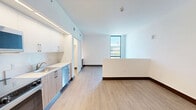 Located at 224 23rd St in Miami Beach, FL, this efficiently designed single-floor unit offers a clean, contemporary aesthetic throughout. The open layout connects the living area and kitchen zones, creating a cohesive and functional space. Two kitchen areas are equipped with stainless steel appliances — including a range, over-the-range microwave, refrigerator, and dishwasher — complemented by white upper cabinetry, warm wood-toned lower cabinets, and light countertops. Under-cabinet lighting adds a modern touch to the kitchen's clean lines. Light wood-look flooring runs continuously throughout the unit, enhancing the sense of openness and warmth. The living area benefits from a large floor-to-ceiling window that brings in ample natural light and provides views of the surrounding Miami Beach neighborhood. A low half-wall partition subtly defines the living space. The hallway provides access to a full bathroom, which features a wood-grain vanity with a white countertop, well-lit by a sleek wall-mounted light fixture. A louvered closet door in the hallway adds a practical storage element. Crisp white walls and dark baseboards frame each room, contributing to the unit's modern, minimalist character throughout.