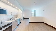 Located at 224 23rd St in Miami Beach, FL, this efficiently designed single-floor unit offers a clean, contemporary aesthetic throughout. The open layout connects the living area and kitchen zones, creating a cohesive and functional space. Two kitchen areas are equipped with stainless steel appliances — including a range, over-the-range microwave, refrigerator, and dishwasher — complemented by white upper cabinetry, warm wood-toned lower cabinets, and light countertops. Under-cabinet lighting adds a modern touch to the kitchen's clean lines. Light wood-look flooring runs continuously throughout the unit, enhancing the sense of openness and warmth. The living area benefits from a large floor-to-ceiling window that brings in ample natural light and provides views of the surrounding Miami Beach neighborhood. A low half-wall partition subtly defines the living space. The hallway provides access to a full bathroom, which features a wood-grain vanity with a white countertop, well-lit by a sleek wall-mounted light fixture. A louvered closet door in the hallway adds a practical storage element. Crisp white walls and dark baseboards frame each room, contributing to the unit's modern, minimalist character throughout.