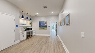 Located at 8710 SW 71st Ave in Ocala, Florida, this well-appointed single-floor commercial space combines modern design with functional versatility. The interior features light wood-look luxury vinyl flooring throughout, crisp white trim, and neutral gray walls that create a clean, professional atmosphere.

A central corridor connects the key areas of the space, lined with curated artwork — including vibrant landscape paintings and botanical prints in elegant frames — adding warmth and visual interest to the circulation areas. Recessed lighting illuminates the hallways, complemented by vaulted ceiling sections that lend an open, airy feel.

The kitchenette area is equipped with white cabinetry, a white subway tile backsplash, a microwave, a beverage refrigerator, and open shelving with greenery accents. Bold blue pendant lights overhead give this area a distinctive, stylish touch.

Two private meeting rooms are accessible from the main corridor, each featuring olive green accent walls, pendant lighting, long work tables, and ergonomic seating — ideal for focused work or small-group sessions. Each room also benefits from natural light through windows. A dedicated conference room is also present, clearly marked with wayfinding signage.

Accessible, gender-neutral restrooms are conveniently located along the main hall. The space also includes safety infrastructure such as a fire extinguisher cabinet and security camera, ensuring a safe and compliant environment.
