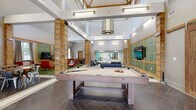 This single-story clubhouse in Lexington, Kentucky, offers a well-appointed community gathering space designed with a contemporary yet inviting aesthetic. The main game room serves as a central focal point, anchored by a rustic wood-framed billiards table set atop herringbone-patterned dark tile flooring. Soaring ceilings with exposed structural elements and decorative wood columns lend an open, airy atmosphere, while a statement pendant chandelier adds a refined touch overhead. Wall-mounted televisions and green turf-accented lounge platforms create distinct activity zones throughout the space.

Adjacent areas include a casual dining zone with round tables and mixed seating, as well as a dedicated card and game table room with tall ceilings, large draped windows, and an arched decorative mirror. A rounded lounge nook features expansive windows overlooking an outdoor pool area, filled with natural light and furnished with a generous sectional sofa. A bar-style kitchenette with a stainless steel counter, wood paneling, and pendant lighting provides a functional refreshment area. The entry corridor features light tile flooring and glass-panel doors leading to a smaller, teal-accented meeting or leasing area, completing this versatile clubhouse layout.