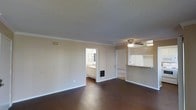 This single-floor space offers a straightforward and functional layout designed for comfortable everyday living. The main living area is finished with rich, dark hardwood-style flooring that contrasts nicely with the light-toned walls, creating a clean and inviting atmosphere. Crown molding runs along the perimeter of the ceiling, adding a touch of classic detail, while a textured ceiling with a ceiling fan provides both character and practical air circulation throughout the room. A pass-through counter connects the living area to the kitchen, promoting an open feel and easy interaction between spaces. The kitchen area features light-colored cabinetry visible through the opening, suggesting ample storage and a bright workspace. A doorway off the main living area leads to what appears to be a bathroom or vanity space, complete with a sink and additional cabinetry, offering convenience and accessibility. A separate door nearby likely leads to a closet or additional room, contributing to the overall utility of the floor plan. The space is well-suited for those seeking a practical, easy-to-maintain layout with consistent finishes throughout.