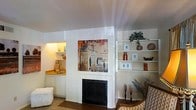 This single-floor space showcases a thoughtfully designed living area centered around a classic fireplace with a clean white surround, serving as the room's focal point. Above the fireplace hangs a bold urban artwork depicting a city skyline, adding a contemporary artistic touch to the otherwise neutral palette. To the right of the fireplace, open built-in shelving spans the wall, offering display space for decorative objects, framed photos, and greenery, contributing to the room's organized yet personalized feel. To the left, a recessed nook houses a compact wet bar or utility counter, complete with cabinetry below and illuminated display shelving above, creating a warm, ambient glow within the space. The walls throughout are finished in crisp white, enhancing the sense of openness and allowing the décor to stand out. The flooring transitions from carpet in the seating area to what appears to be hardwood or laminate near the fireplace. Candlestick holders and decorative botanicals add warmth and texture, while patterned upholstered seating contributes to the inviting, lived-in atmosphere of this well-appointed single-level home.