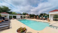 This inviting outdoor amenity space centers around a generously sized, crystal-clear swimming pool with a smooth concrete surround that provides ample room for lounging and recreation. The pool area is thoughtfully designed with multiple seating arrangements, including cushioned lounge chairs and chairs with vibrant orange accents, offering comfortable spaces for relaxation. A classic white pergola with decorative columns anchors one side of the pool deck, providing shaded respite, while a charming brick-accented gazebo adds architectural interest and additional covered seating nearby. Decorative planters with greenery accent the pool surround, adding a touch of natural beauty to the hardscaped area. To one side, a colorful children's playground equipped with climbing structures, slides, and activity panels offers a dedicated recreational space for younger residents. The entire area is framed by mature trees and lush landscaping, creating a sense of privacy and a serene, park-like atmosphere. Tall light poles are positioned throughout the space, ensuring the amenity area remains functional and welcoming into the evening hours. Overall, the space delivers a resort-inspired outdoor experience combining relaxation, recreation, and natural surroundings.