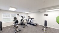 Located at 907 Ikes Peak Rd in Clinton, Iowa, this dedicated exercise room offers a compact yet efficiently organized fitness space. The room features clean white walls, textured grey carpet flooring, and flush-mounted panel lighting that ensures the space is consistently well-lit. Natural light filters in through multiple windows fitted with white blinds, adding to the bright and inviting atmosphere.

The cardio equipment selection is well-rounded, including a treadmill, an elliptical trainer, a recumbent bike, and an upright stationary bike — providing a variety of low- and high-impact workout options. For strength and functional training, the room is equipped with a tiered dumbbell rack, an adjustable workout bench, resistance bands, medicine balls in assorted sizes, and exercise balls. A Smart Fitness accessory station rounds out the functional training corner.

Motivational fitness artwork is displayed on the walls, contributing to an energizing environment. A hand sanitizer station and waste bin are also present, reflecting attention to cleanliness and user convenience. The space is thoughtfully arranged to maximize floor space and allow comfortable movement between equipment, making it a practical and complete fitness facility.