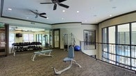 This fitness center offers a thoughtfully designed workout space that combines functionality with an open, airy atmosphere. The single-floor layout is finished with durable carpet flooring and features crown molding details along the upper walls, giving the space a polished appearance. Ceiling fans are installed throughout, ensuring consistent air circulation during workouts.

The facility is well-lit through a combination of recessed lighting and large windows that invite natural light while providing views of the surrounding area. A curved metal railing defines an open boundary along one side of the space, adding an architectural element that enhances the room's flow.

Equipment is thoughtfully arranged throughout the floor. A dedicated free weights area includes a full rack of dumbbells and flat workout benches for strength training. A multi-functional cable machine occupies the center of the room, accommodating a wide range of exercises. An adjustable workout chair and additional benches provide versatility for various routines, while a stability ball rounds out the available equipment.

Full-length mirrors are positioned along one wall to assist with form and technique, and glass doors lead to adjacent areas, suggesting additional amenities nearby. Overall, this fitness center provides a comprehensive and well-maintained environment suitable for a range of workout needs.