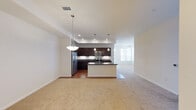This single-floor residence showcases a thoughtfully designed open-concept layout that seamlessly integrates the kitchen and living areas into one cohesive space. The kitchen is a standout feature, appointed with rich dark wood cabinetry, sleek dark granite countertops, and a full suite of stainless steel appliances, including a French door refrigerator with ice and water dispenser, an electric range, and an over-the-range microwave. A peninsula with an undermount sink and breakfast bar extends into the living space, providing both additional prep space and a casual dining area. Pendant lighting hangs elegantly above both the peninsula and the adjacent living area, complementing the recessed lighting throughout to create a warm and well-lit atmosphere. The kitchen features hardwood flooring that transitions cleanly into plush wall-to-wall carpeting in the living area, adding warmth and comfort underfoot. Crisp white walls throughout the space enhance the sense of openness and brightness. The overall design balances modern finishes with functional layouts, making it a practical and stylish living environment.
