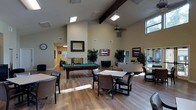 This single-floor recreational space is designed to serve as a multifunctional gathering and entertainment hub. The room's most striking architectural feature is its soaring vaulted ceiling, accented by prominent dark wood beams that add a rustic yet refined character. Skylights and high clerestory windows allow generous natural light to flood the interior, complementing the warm-toned hardwood-style flooring throughout.

A full-size pool table serves as a central focal point, positioned beneath a pendant light fixture that draws the eye upward toward the dramatic ceiling height. Adjacent to it, a wall-mounted flat-screen television is flanked by lush potted plants, adding a touch of greenery to the neutral, beige-toned walls. A fireplace with a decorative tile surround and a large mirror above it anchors one side of the room, lending warmth and elegance.

The space is thoughtfully zoned, featuring a comfortable lounge area with sofas and chairs, as well as smaller dining and activity tables with upholstered seating. Views of a bright outdoor area are visible through large windows and a doorway, enhancing the open, airy ambiance of the space.