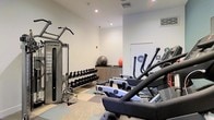 This fitness center offers a comprehensive workout environment designed to accommodate a range of exercise routines. The space features a clean, modern aesthetic with white walls, recessed lighting, and a multi-tone tile floor that adds visual interest underfoot. Professional-grade Cybex strength training equipment anchors one side of the room, complemented by a full dumbbell rack with weights neatly organized in ascending order for easy access. Cardio equipment, including elliptical machines, is positioned to allow ample space for movement between stations. A low wooden storage console along the back wall holds exercise balls, including a stability ball and medicine ball, providing options for functional and core-focused training. Restroom facilities are conveniently accessible directly from the fitness area, as indicated by clearly marked signage near the rear door. The recessed ceiling lights provide consistent, even illumination throughout the space, creating a bright and energizing atmosphere. The layout is thoughtfully organized to maximize usability while maintaining an uncluttered, functional environment suited for a focused fitness experience.