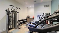 This fitness center offers a comprehensive workout environment designed to accommodate a range of exercise routines. The space features a clean, modern aesthetic with white walls, recessed lighting, and a multi-tone tile floor that adds visual interest underfoot. Professional-grade Cybex strength training equipment anchors one side of the room, complemented by a full dumbbell rack with weights neatly organized in ascending order for easy access. Cardio equipment, including elliptical machines, is positioned to allow ample space for movement between stations. A low wooden storage console along the back wall holds exercise balls, including a stability ball and medicine ball, providing options for functional and core-focused training. Restroom facilities are conveniently accessible directly from the fitness area, as indicated by clearly marked signage near the rear door. The recessed ceiling lights provide consistent, even illumination throughout the space, creating a bright and energizing atmosphere. The layout is thoughtfully organized to maximize usability while maintaining an uncluttered, functional environment suited for a focused fitness experience.