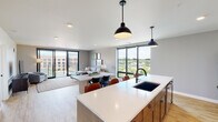This contemporary single-floor residence is designed with an open-concept layout that seamlessly connects the kitchen, dining, and living areas into one cohesive, light-filled space. Large floor-to-ceiling windows with sleek black frames flood the interior with natural light and provide expansive views of the surrounding greenery and neighboring architecture. The kitchen is outfitted with white shaker-style cabinetry, a white subway tile backsplash, stainless steel appliances — including a refrigerator, range, and microwave — and a generously sized island with a white quartz countertop, undermount black sink, and pendant lighting. Light wood-tone flooring flows throughout the space, adding warmth to the modern palette. The living area opens directly onto a private balcony through sliding glass doors, offering an inviting outdoor retreat. The bedroom is a calm, understated space with a large window, a built-in closet with dual hanging rods, and additional storage options. A clean, neutral bathroom features a walk-in shower, tile flooring, and a vanity with a white countertop. The in-unit laundry room houses a full-size washer and dryer alongside a water heater and HVAC system, providing complete functionality within the residence.
