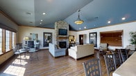 This single-story clubhouse at Fresh Wind Rentals presents an inviting community gathering space defined by thoughtful architectural details and quality finishes throughout. The exterior makes a strong first impression with a stacked stone façade, a covered entryway supported by white columns, and a rich wood-framed glass door, all framed by colorful landscaping and ornamental grasses.

Inside, the main lounge area is anchored by a striking floor-to-ceiling stacked stone fireplace with a mounted flat-screen TV above the mantle. Soaring vaulted ceilings painted in a cool blue-gray tone add a sense of openness and height, while recessed lighting and a decorative pendant light fixture provide warm, layered illumination. Rich hardwood flooring flows throughout the space, complementing the neutral furnishings that include plush sofas and accent seating arrangements. Large windows allow natural light to filter in, and additional seating areas with bistro-style tables offer flexible use of the space.

The exterior rear of the building opens to a generously sized outdoor swimming pool with a clean concrete surround. A pergola-shaded patio with wrought iron table-and-chair sets provides an ideal outdoor retreat, all enclosed by a black iron fence with open views of the surrounding landscape and sky.