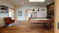 This single-floor space masterfully combines rustic warmth with contemporary design across a series of open, interconnected living areas. The gourmet kitchen is a true centerpiece, boasting soaring vaulted ceilings with exposed timber beams and expansive floor-to-ceiling windows that flood the space with natural light. A large island with a reclaimed wood base and crisp white quartz countertop anchors the room, surrounded by industrial-style brass barstools. Soft grey cabinetry, white subway tile backsplash, a stainless steel range hood, and oversized brass pendant lights complete the refined yet rugged aesthetic. Large-format grey tile flooring grounds the space. Adjacent to the kitchen, the dining area transitions to rich wide-plank hardwood floors and features high ceilings with elegant gold pendant lighting. A built-in sideboard with a wine rack, open shelving, and a marble-topped counter lines a warm earth-toned accent wall. The entertainment area is equally impressive, offering a full-size billiards table beneath sleek pendant lights and a skylight above. Beautifully crafted sliding barn doors with etched glass panels connect the spaces, while tufted leather armchairs provide a comfortable lounge setting. The cohesive design language throughout makes this a sophisticated and inviting space.