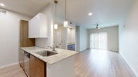 This single-floor residence showcases a cohesive modern design with light wood-tone flooring running throughout the main living areas, complemented by neutral walls that create a bright, airy atmosphere. The galley-style kitchen is a standout feature, outfitted with two-tone cabinetry pairing flat-front white upper cabinets with warm wood-grain lower cabinets. Light granite countertops provide ample prep space, and stainless steel appliances, including a refrigerator and range, complete the functional layout. Pendant lighting over the peninsula adds a stylish touch.

The open living area extends from the kitchen, bathed in natural light from a large sliding glass door fitted with vertical blinds. A ceiling fan overhead adds comfort to the space. A secondary room features the same light hardwood flooring and another large window that fills the area with natural light. The bedroom is carpeted for warmth and comfort, with its own ceiling fan and a window offering outside views.

The bathroom maintains the design language of the unit, featuring a wood-grain vanity with a white countertop, a large mirror, a tiled shower-tub combination, and light wood-look flooring. A hallway connects the living areas to the bathroom, with in-unit laundry access nearby, rounding out this well-appointed residence.
