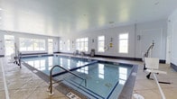 Located at 700 Citation Lane in Cherry Hill, New Jersey, this single-floor indoor aquatic facility offers a clean, well-maintained environment designed for both recreation and relaxation. The centerpiece of the space is a generously sized indoor swimming pool with a classic blue tile border and clearly marked depth indicators, providing a safe and inviting environment for swimmers of all levels. Adjacent to the main pool is a separate hot tub or spa area, distinguished by its own entry railings and hydrotherapy jets visible beneath the water's surface. The surrounding pool deck is finished in neutral-toned tile and is furnished with reclining lounge chairs, offering a comfortable area to rest before or after a swim. The interior is bright and airy, thanks to a series of large, evenly spaced windows along the perimeter walls that allow abundant natural light to fill the space and provide views of the outdoor greenery beyond. The walls are finished in a crisp white, complemented by a smooth, light-toned ceiling that enhances the open, spacious feel of the room. Safety equipment, including a life ring, is visibly mounted on the wall, reflecting a commitment to facility safety standards. Overall, the space presents a polished, functional, and welcoming aquatic environment.