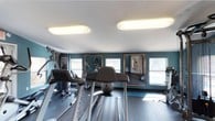 This fitness center is a thoughtfully designed exercise space that offers everything needed for a comprehensive workout. The room is finished with bold teal-blue accent walls that create an energizing atmosphere, complemented by a clean white ceiling with modern flush-mount lighting panels that ensure bright, even illumination throughout. Large windows along the walls flood the space with natural light, enhancing the open and airy feel of the room. A series of framed wall mirrors are strategically positioned to visually expand the space and allow exercisers to monitor their form during workouts. The flooring features a durable, fitness-appropriate surface well-suited for high-traffic use. The space is outfitted with a selection of Matrix-brand cardio equipment, including treadmills and a stationary bike, as well as a multi-function strength training machine and free weight accessories such as a medicine ball. The layout is organized to allow comfortable movement between stations. Overall, this fitness center strikes a practical and motivating balance between functionality and design, making it an inviting space for a wide range of exercise routines.