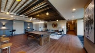 Located at 265 E State St in the heart of Columbus, Ohio, this contemporary amenity lounge is a thoughtfully designed communal space that blends upscale aesthetics with functional social areas. The open floor plan is anchored by wide-plank hardwood flooring that flows seamlessly throughout, connecting each distinct zone of the space.

A central billiards area features a full-size pool table beneath a dramatic exposed beam ceiling and a cluster of elegant pendant lights, setting an inviting, lounge-like atmosphere. Adjacent to the billiards zone is a relaxed seating area appointed with a plush gray sectional, curated accent chairs, and a decorative sunburst mirror. A separate lounge nook showcases a bold navy velvet sofa, textured accent chairs, and framed art, complemented by a tray ceiling with recessed lighting.

The space also includes a dedicated bar and kitchenette area outfitted with a long white quartz island, bar stools, under-counter refrigeration, a sink, and a mounted flat-screen TV beneath a textured tile backsplash. A cozy fireplace lounge with golden velvet sofas and a moody, dark accent wall with sconce lighting provides an intimate retreat within the larger space.

Floor-to-ceiling glass doors open to a furnished outdoor patio with umbrella-shaded dining tables and string lights, offering a seamless indoor-outdoor experience in the heart of downtown Columbus.