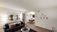 This single-floor space offers a thoughtfully designed open-concept layout that seamlessly connects the living and dining areas. The living room is anchored by a sleek dark sofa complemented by an assortment of accent pillows, a round glass coffee table, and a plush area rug, creating a welcoming and stylish atmosphere. Framed artwork adorns the crisp white walls, adding character to the space alongside decorative circular mirrors. Warm-toned, wood-look flooring flows throughout, tying the areas together cohesively. A floor lamp provides supplemental lighting, enhancing the room's bright and airy feel. An open passageway leads directly into the dining area, which features a compact dining table surrounded by modern chairs, ideal for casual meals. A pendant light fixture hangs above, providing focused illumination. The dining space is further accented by a chalkboard wall feature, lending a playful and personalized touch. The overall color palette is neutral and clean, dominated by white walls and warm flooring tones, creating a balanced and inviting ambiance throughout the home.