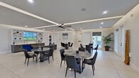 This contemporary single-floor space is designed as a modern communal gathering area, combining elegance with functional versatility. The expansive main hall features large-format porcelain tile flooring throughout, crisp white walls, and a coffered tray ceiling accented by recessed LED lighting and integrated cove illumination that lends a warm, polished ambiance. A large-blade ceiling fan anchors the space, ensuring comfortable airflow across the generous open floor plan.

Multiple round marble-top dining tables, each paired with sleek dark upholstered chairs, are arranged throughout the hall to accommodate groups of varying sizes. A lounge area is furnished with a plush tufted velvet sofa and accent chair in rich blue and grey tones, adding a sophisticated social setting. Built-in shelving along one wall displays curated décor accents alongside a large scenic display screen, while tall potted palms introduce natural warmth to the interior.

Floor-to-ceiling glass doors and windows on multiple sides flood the space with natural light and provide views of the surrounding greenery and outdoor areas. A separate, fully equipped kitchen features grey flat-panel cabinetry, a mosaic tile backsplash, stainless steel appliances including a gas range, microwave, and a large side-by-side refrigerator, with ample counter space for catering and event preparation.