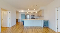 Located at 3514 Aspire Cir in Cape Coral, Florida, this single-floor residence presents a clean, contemporary design characterized by light wood flooring, crisp white walls, and an open layout that promotes a bright and airy atmosphere throughout. The open-concept kitchen serves as a central feature of the living space, anchored by a large peninsula with a light-colored countertop that provides ample workspace and casual seating potential. The kitchen is outfitted with stainless steel appliances, including a refrigerator, range, and built-in microwave, complemented by soft-toned cabinetry with modern hardware and a stainless steel undermount sink. Pendant lighting over the peninsula adds warmth, while a beverage cooler offers a practical touch. The living area is generously proportioned, with a ceiling fan overhead and full-height sliding glass doors dressed in vertical blinds that flood the space with natural light and provide outdoor access. A hallway connects the living areas to the bathroom, which features a clean walk-in shower with glass enclosure and a light stone-topped vanity with an undermount sink. The overall design aesthetic is modern and minimalist, well-suited to the sunny Florida lifestyle.