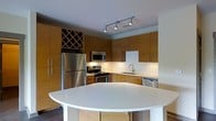 This single-floor space showcases a thoughtfully designed interior with a strong emphasis on modern aesthetics and functional living. The kitchen serves as a focal point, featuring a full suite of stainless steel appliances, including a refrigerator, electric range with microwave above, and dishwasher. Warm, flat-panel wood cabinetry lines the walls from floor to ceiling, offering generous storage throughout. A distinctive diamond-pattern wine rack built into the upper cabinetry adds a refined, custom touch. Under-cabinet lighting illuminates the clean white countertops and tile backsplash, enhancing the sense of brightness in the space. A curved, white kitchen island with bar seating extends outward, creating a natural divide between the kitchen and the living area while providing additional workspace and casual dining options. Track lighting on the ceiling offers directed, adjustable illumination across the kitchen. The dark hardwood flooring grounds the space with contrast against the lighter cabinetry and countertops. Overall, the space conveys a clean, contemporary feel with well-chosen materials and a practical layout suited for everyday living.