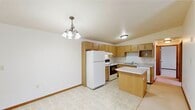 This single-floor unit offers a practical and efficient layout across three bedrooms and two bathrooms. The open kitchen serves as a central hub, featuring warm oak cabinetry extending to the ceiling for ample storage, light-toned countertops, a peninsula island for additional workspace or casual dining, and white appliances including a refrigerator and electric range. Light tile flooring runs through the kitchen and entry areas, contributing to a clean, easy-maintenance aesthetic. A decorative chandelier accents the dining area adjacent to the kitchen.

The living room is carpeted and opens directly to a private balcony through a sliding glass door, providing outdoor views and natural light. A baseboard heating system lines the living room wall. The bedrooms are carpeted and feature neutral white walls with warm wood baseboards. The primary bedroom benefits from a dedicated walk-in closet equipped with wire shelving and hanging rods for organized storage.

The bathrooms include a full tub-and-shower combination with clean white surround panels. Throughout the unit, warm wood-toned doors and trim provide consistent detailing, while the neutral color palette creates a cohesive, functional living environment.