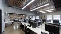 This computer lab combines industrial character with modern functionality in a single open-plan floor. The space is defined by its striking exposed brick walls and a warm wood-paneled ceiling, which contrast beautifully with the clean, white workstation surfaces arranged in organized rows throughout the room. Overhead, rows of bright fluorescent strip lighting ensure consistent, even illumination across all work areas, supplemented by natural light streaming in through multiple windows fitted with adjustable blinds along the perimeter walls.

Each workstation is equipped with a desktop computer tower, monitor, keyboard, and ergonomic task chair, accommodating a large number of users simultaneously. The workstations are arranged in clusters and rows, promoting an efficient use of the open floor plan while allowing comfortable movement between stations. The polished concrete floor adds to the industrial aesthetic and is easy to maintain.

The high ceiling height lends the lab an open, airy atmosphere, reducing the sense of crowding even when fully occupied. Additional wall-mounted air conditioning units support climate control throughout the space. Decorative artwork on the walls adds a touch of personality, making this computer lab both a practical and visually engaging environment for work or study.