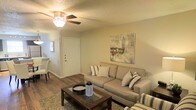 This single-floor space presents a well-appointed living area designed with both comfort and functionality in mind. The living room serves as the focal point, featuring rich hardwood-style flooring that contrasts tastefully with the soft, neutral-toned walls. A ceiling fan with an integrated light fixture is centrally mounted, providing both air circulation and ambient lighting throughout the space.

The living area is anchored by a plush, beige sectional sofa accented with striped throw pillows, complemented by a dark wood coffee table that adds a grounded, earthy element to the room. A framed cityscape wall print adds a decorative touch and visual interest to the space. A textured area rug beneath the seating arrangement helps define the living zone, adding warmth underfoot.

A clear sightline from the living room leads to the entryway, where a solid white front door is visible, flanked by a glimpse of an adjacent area that appears to offer additional living or dining space. The textured ceiling adds architectural character, while the overall layout promotes an open, easy flow between areas, resulting in a welcoming and practical living environment.