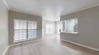 This single-floor residence offers a clean, modern aesthetic defined by light neutral tones, light wood-look flooring, and crisp white trim throughout. The open-concept living and dining area is generously proportioned, filled with natural light through large windows dressed with white horizontal blinds. Crown molding adds a refined architectural detail to the living space, which flows seamlessly into the kitchen via a peninsula countertop with an undermount sink — ideal for casual dining or meal prep interaction. The kitchen is well-appointed with gray shaker-style cabinetry, white countertops, a classic subway tile backsplash, and a full suite of stainless steel appliances including a refrigerator, range, and microwave. A dedicated entryway with a thermostat panel and overhead HVAC vent provides a practical transition into the home. The bedrooms are spacious and bright, each benefiting from the same light flooring and neutral wall palette, with ample wall space and closet access. The bathroom features a framed vanity mirror with warm metallic trim, a sleek white quartz countertop, undermount sink, modern chrome faucet, and gray cabinetry with brushed hardware — completing the home's cohesive, contemporary design language.